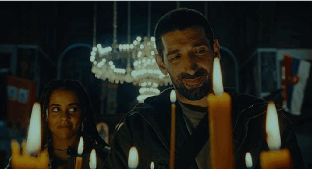 Still from a film showing a man and women in a dark room with a chandelier behind them and candles in front. The man is smiling as he looks at the candles; the woman is looking across at him with a hint of a smile.
